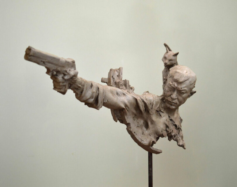 A clay sculpture by Neil Ferrier depicts a man aiming a pistol with an intense expression; a small cat sits on his shoulder. The piece is unfinished and partially broken.