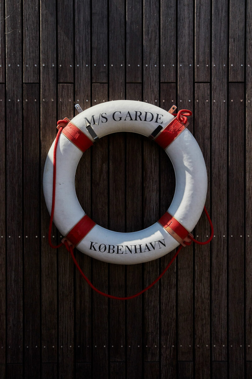 A white lifebuoy with red bands and the text "M/S GARDE KØBENHAVN" is mounted on a dark wooden wall.