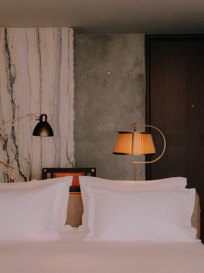 A neatly made bed with white pillows and sheets, two lamps on either side, and a marble and concrete accent wall in the background.