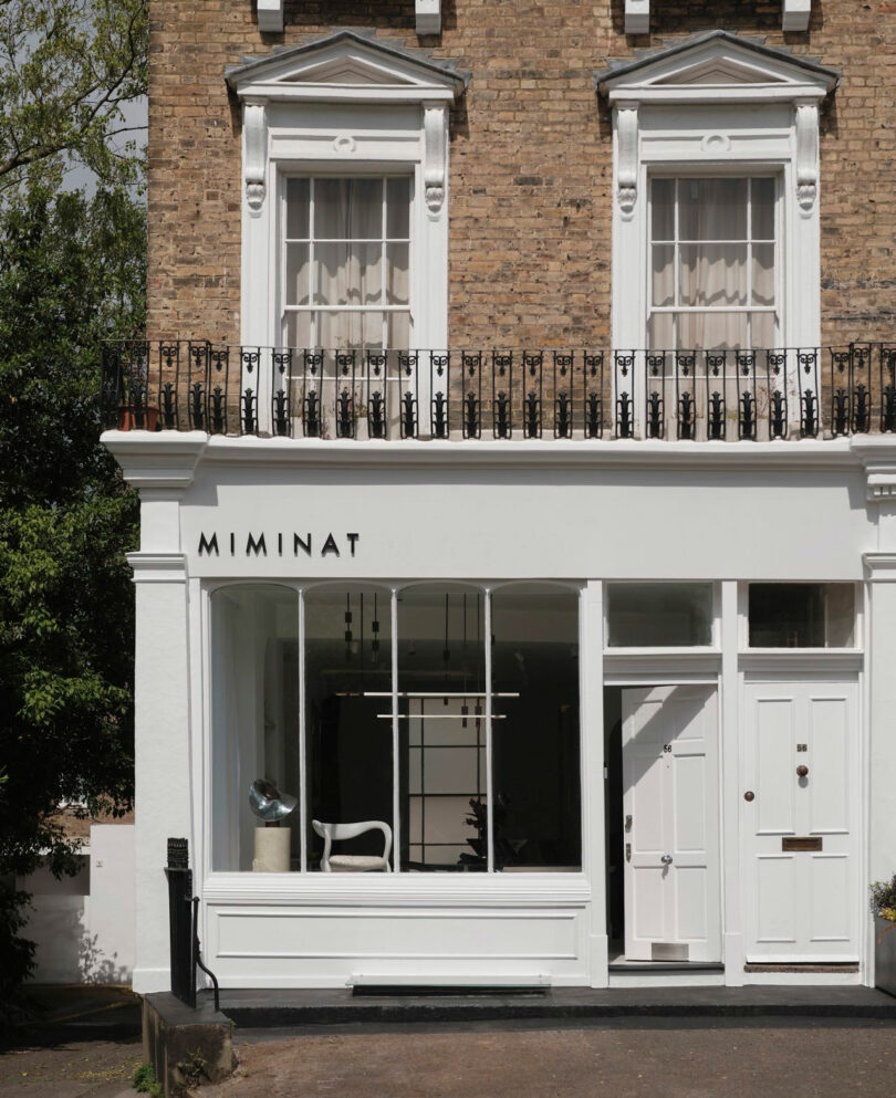 The storefront of Miminat features large display windows, a pristine white façade, and a decorative brick upper level with two windows and elegant balcony railings.