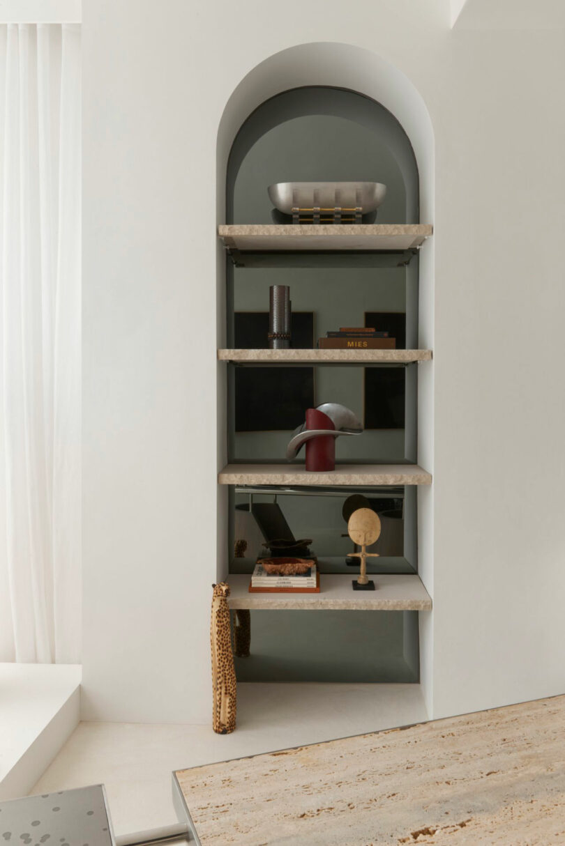 A built-in Miminat arched shelving unit with four stone shelves displays decorative objects, including books, a bowl, a small sculpture, and a globe, set against a neutral-toned wall.