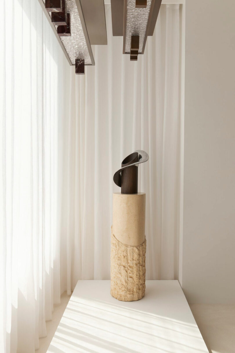A modern abstract sculpture by Miminat, with metal and wood elements, stands on a cylindrical stone pedestal in a minimal, light-filled room with sheer white curtains.