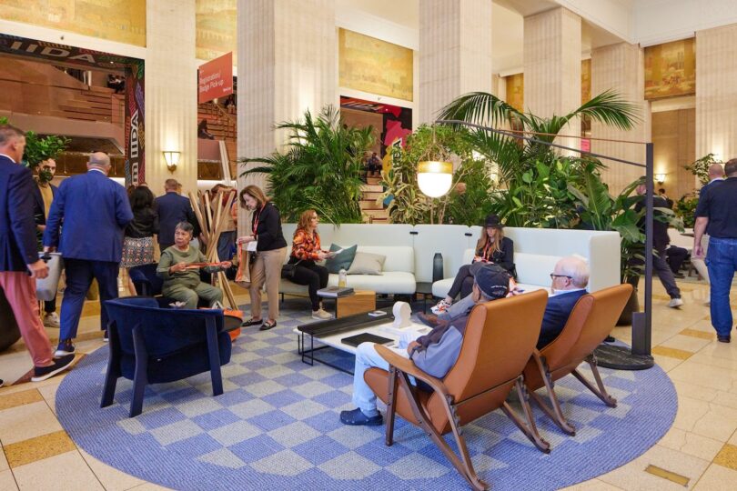 People are sitting and standing in a modern, indoor lounge area with plants, sofas, and armchairs, while others pass by or converse in the background