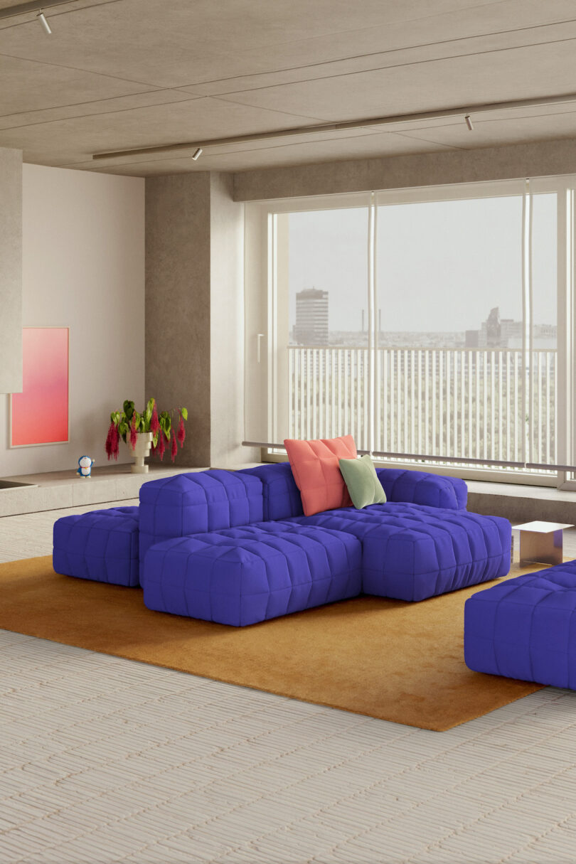 Modern living room with large windows, a blue modular sofa, pink and green cushions, brown rug, potted plant, and cityscape view in the background