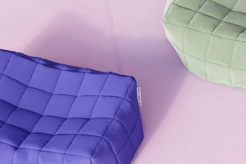 A quilted purple cushion and a quilted light green cushion are placed on a smooth, light pink surface