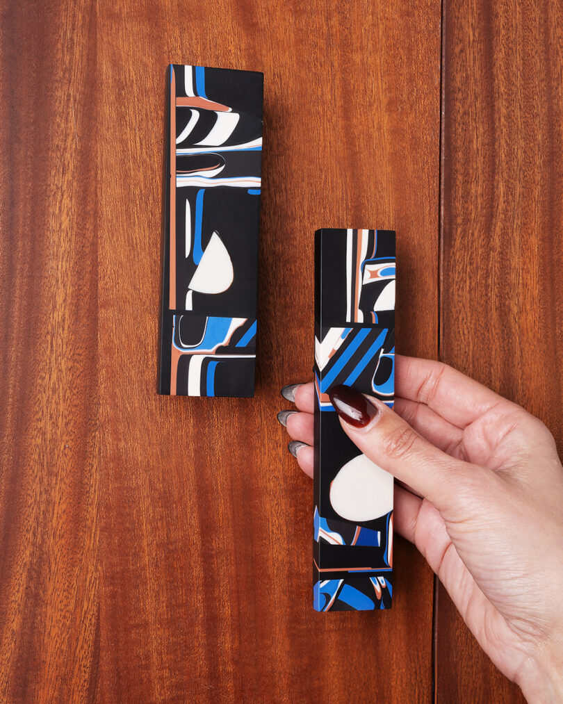 A hand holds a rectangular, abstract-patterned box with black, white, blue, and red details; a second identical box rests on a wooden surface