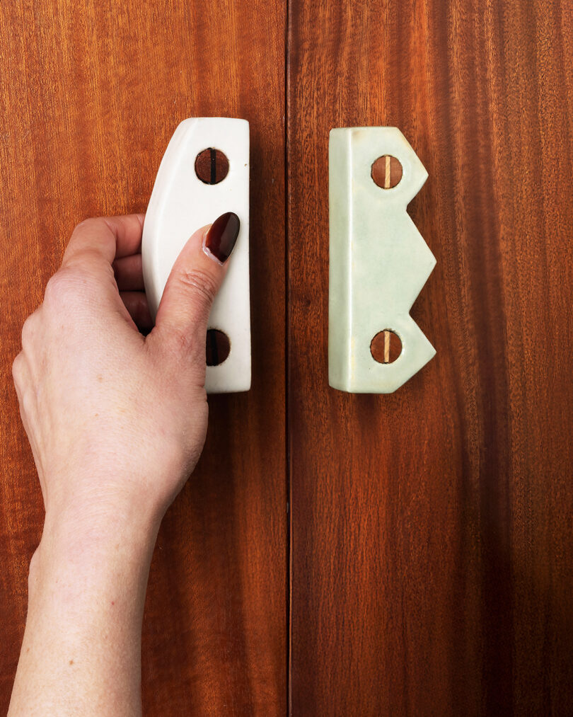 A hand with painted nails holds a white ceramic handle on a wooden cabinet, with a matching but differently shaped handle on the right door