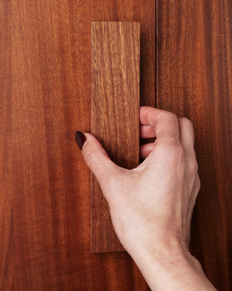 A hand with painted nails holds a vertical wooden handle attached to a wooden surface, likely a cabinet or door