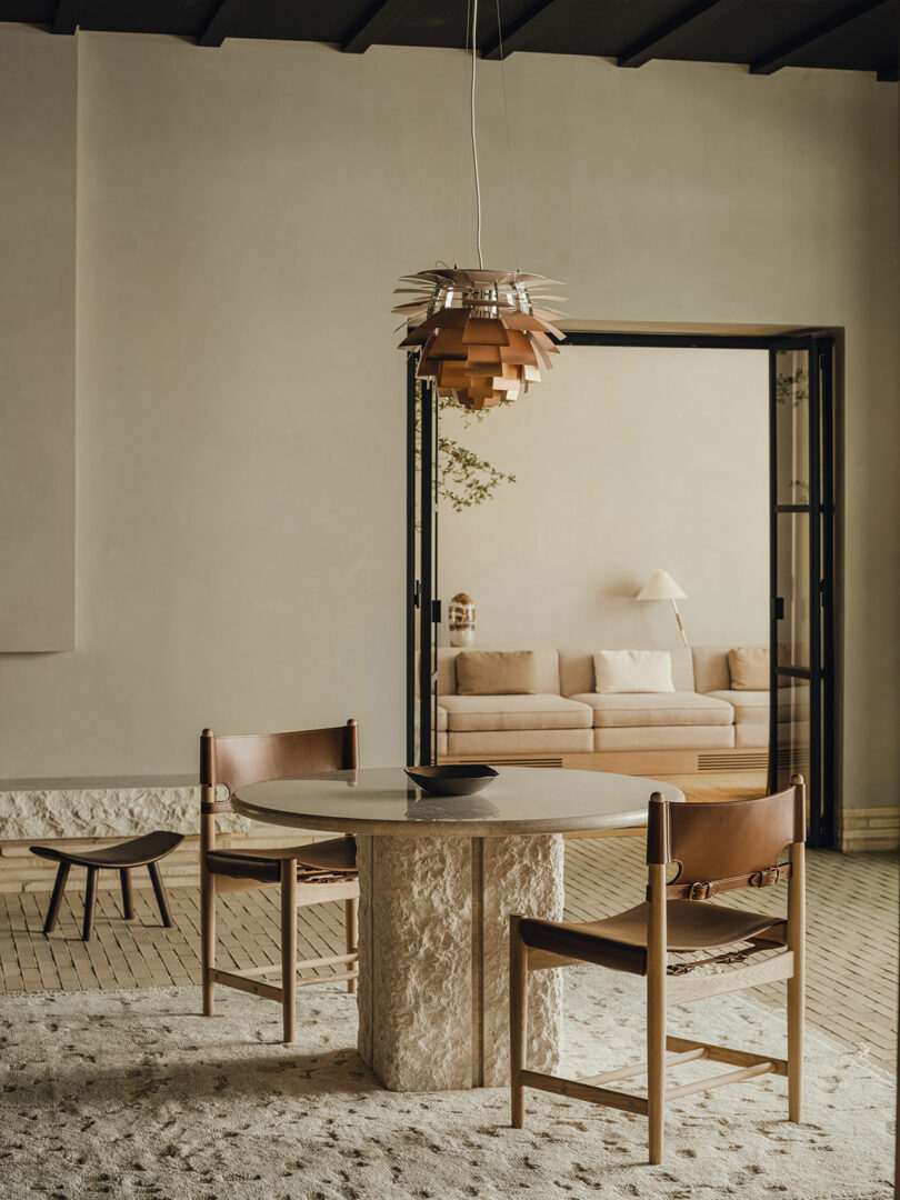 A minimalist dining area with a round stone table, two wooden chairs with brown leather seats, and a modern pendant light, opening to a neutral-toned living room with a sofa and lamp.