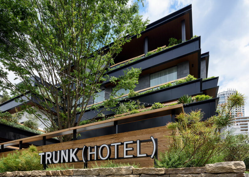 A modern multi-story hotel building with layered balconies and greenery, with a sign reading "TRUNK (HOTEL)" in the foreground.