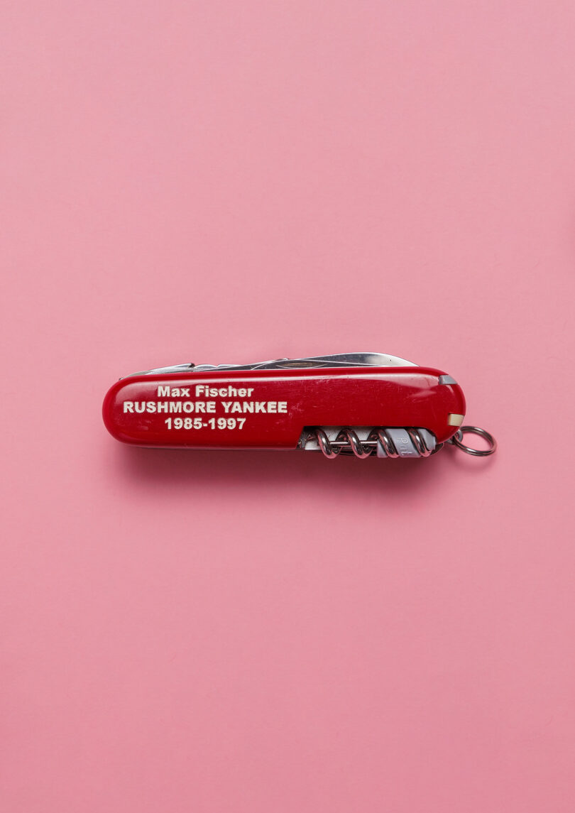 A red Swiss Army knife engraved with “Max Fischer RUSHMORE YANKEE 1985-1997” lying on a pink background.