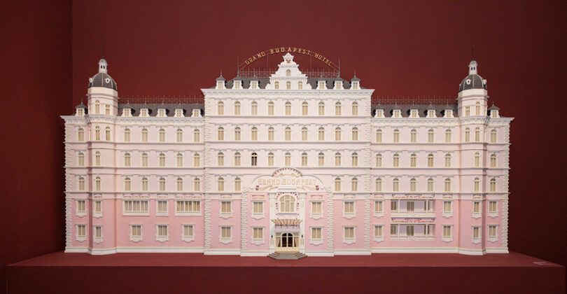 A symmetrical, detailed model of the Grand Budapest Hotel with pink and white facade set against a deep red background.